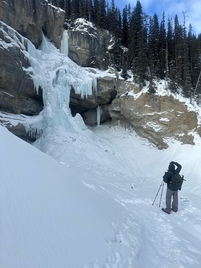 A photographer with a tripod standing in a snowy basin below a large frozen waterfall on a grey cliff.