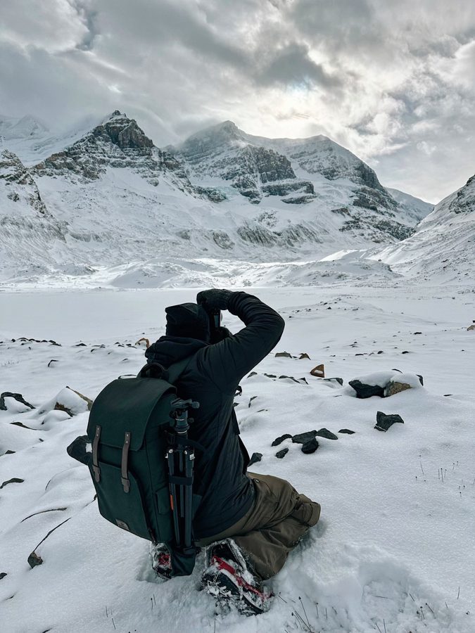 A photographer kneeling in deep snow, camera raised toward a glaciated mountain range under heavy cloud.