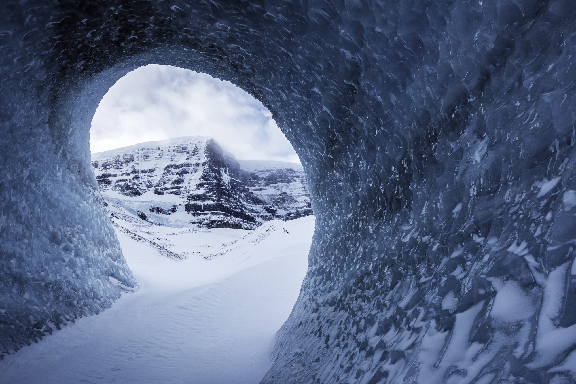 Looking out of a wind carved ice tunnel through a round opening to a snowy peak under cloud.