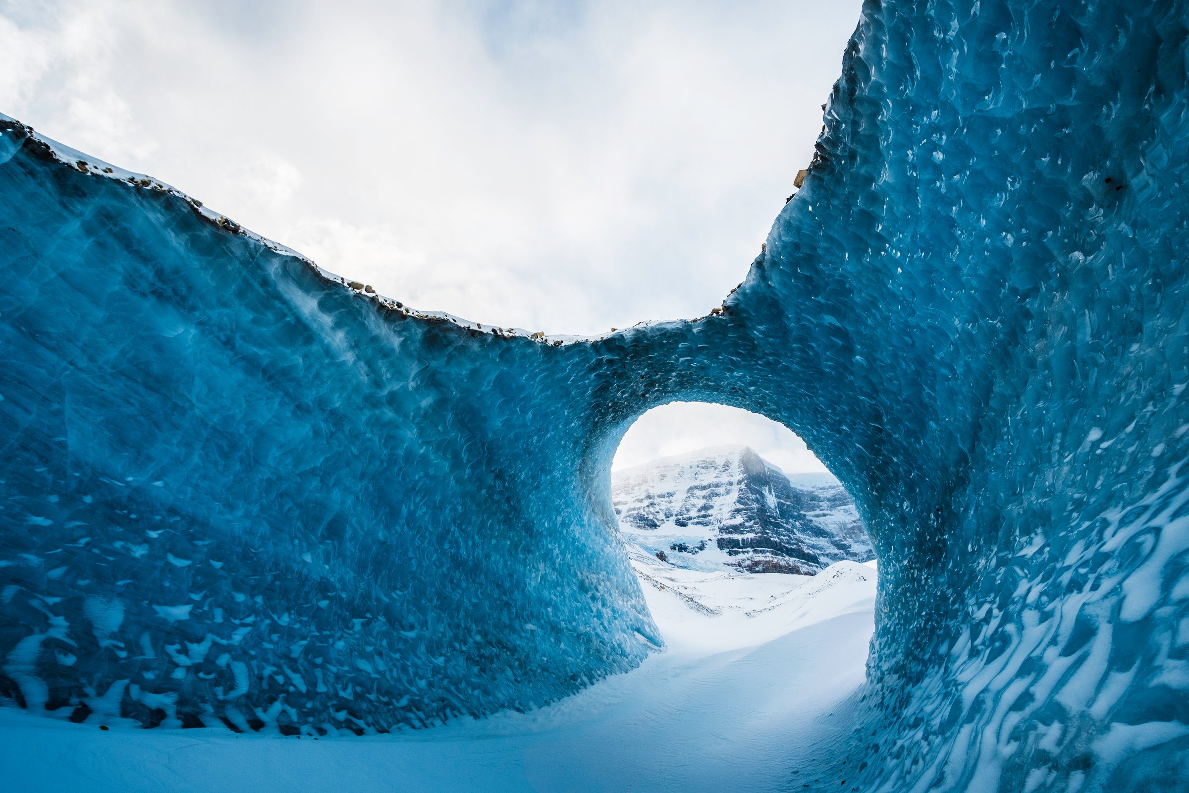 A wave shaped arch of blue ice framing a small round opening, with a snowy peak beyond.