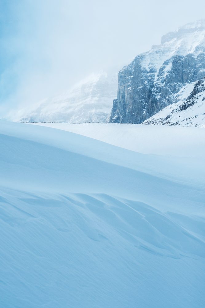Wind shaped snow drifts filling the foreground, with a cliff wall fading into mist.