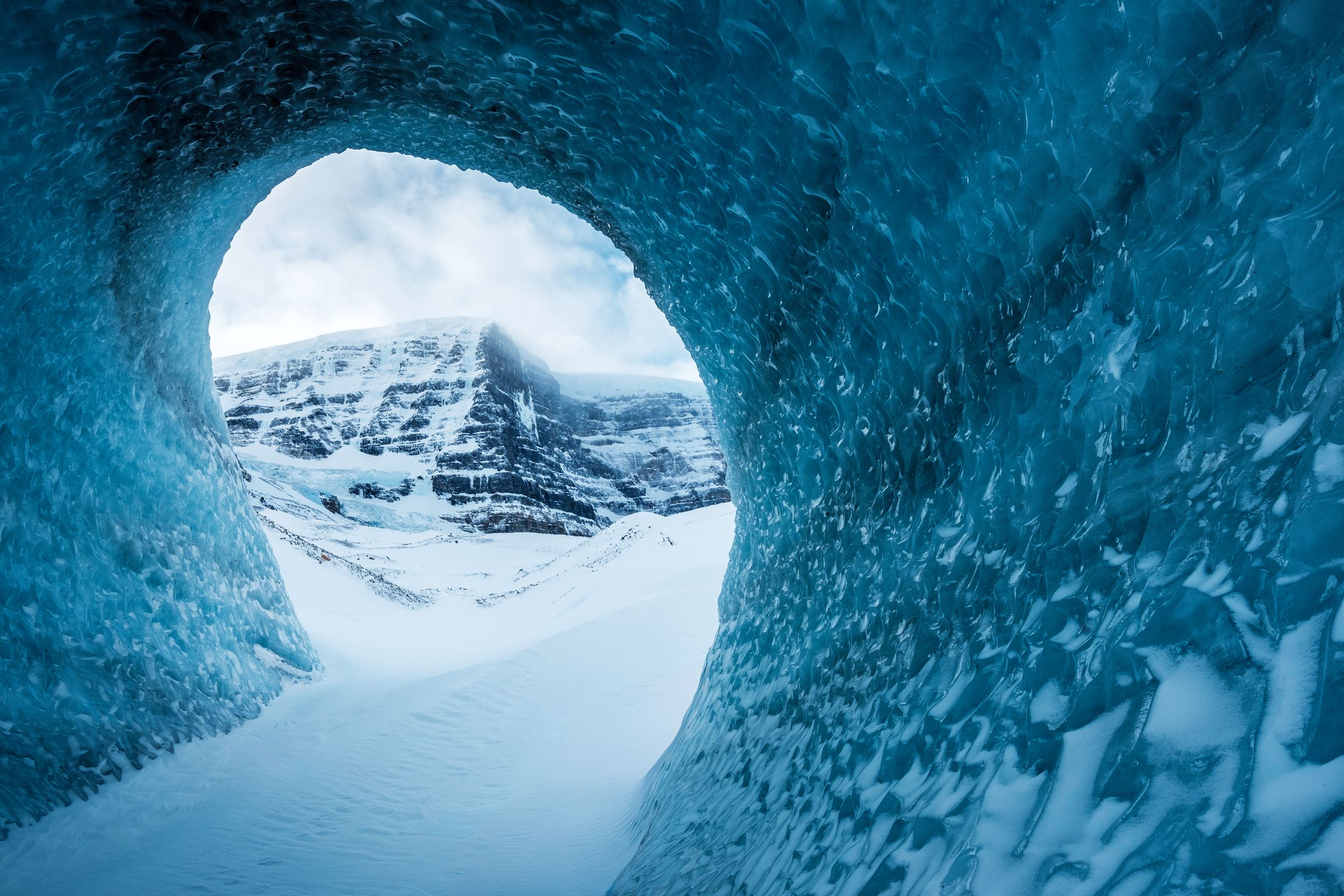 Looking out through a round opening in a wind carved ice tunnel to a snowy peak under cloud.