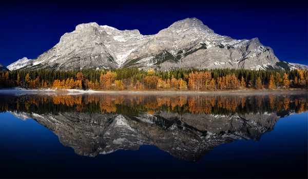 A jagged range and a strip of orange autumn trees mirrored in a still pond.