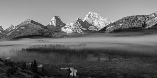 Black and white wide range with a lit peak, mist through the forest and a river across the valley floor.
