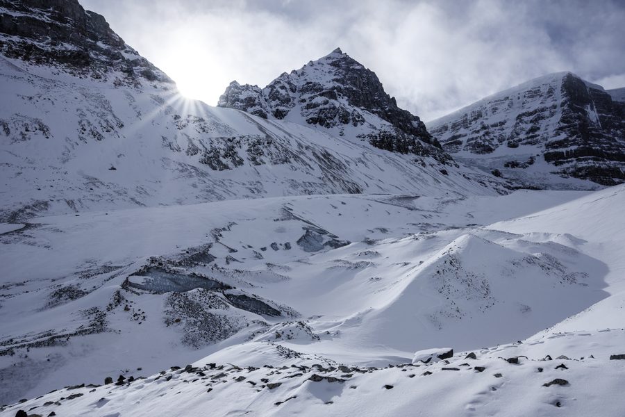 Low sun bursting over a snowy ridge, with blue pools of ice scattered across the glacier.