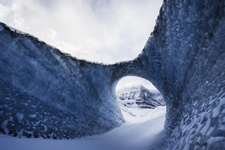 A wave shaped arch of blue ice framing a small round opening, with a snowy peak beyond.