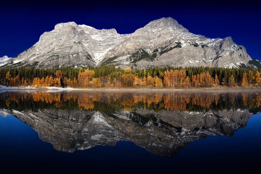 A grey limestone range and a band of autumn gold trees mirrored in perfectly still water under a deep blue sky.