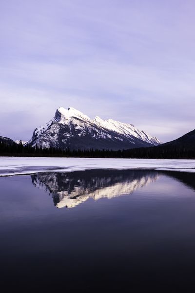 Snow covered peak with a lit summit reflected in dark open water beside a shelf of white ice, under a pale lavender sky.