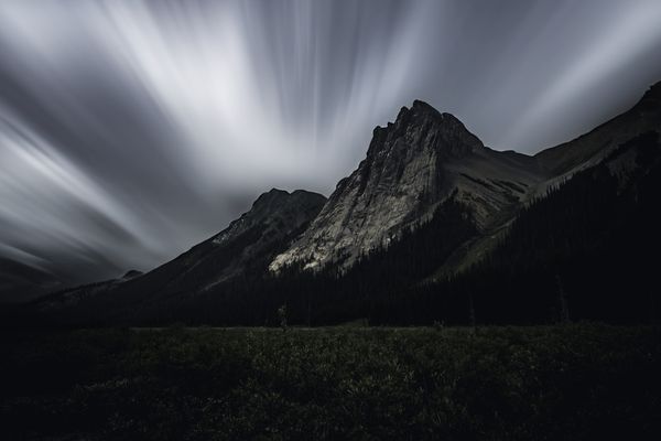 Jagged rocky peak over a dark forest with long exposure clouds streaking across the sky.